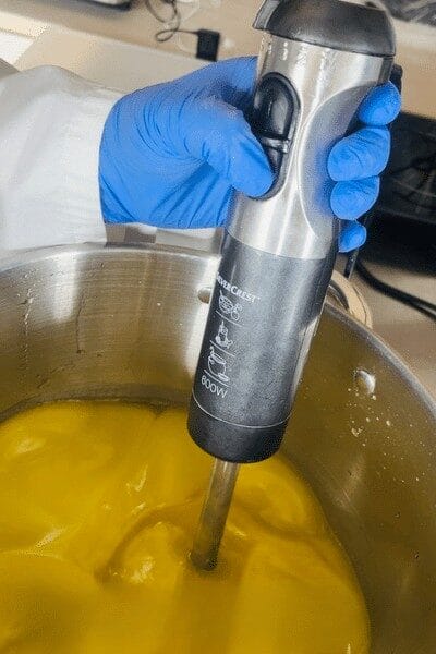 Une main gantée utilise un mixeur plongeant pour mélanger une préparation jaune dans une grande marmite en inox.